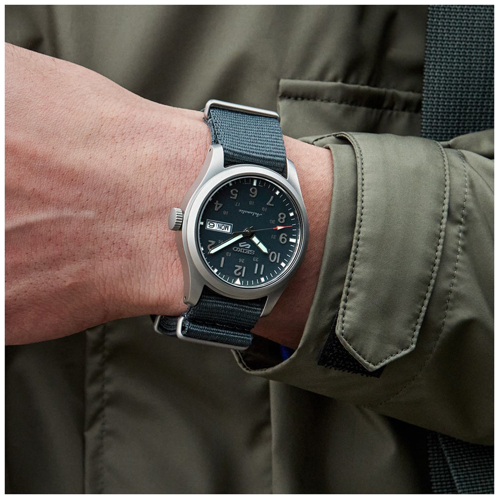 Een persoon in een donkergroen jasje toont het Seiko 5 Sports herenhorloge SRPG31K1 van Seiko, met een donkere wijzerplaat en stoffen band met dag- en datumweergave.