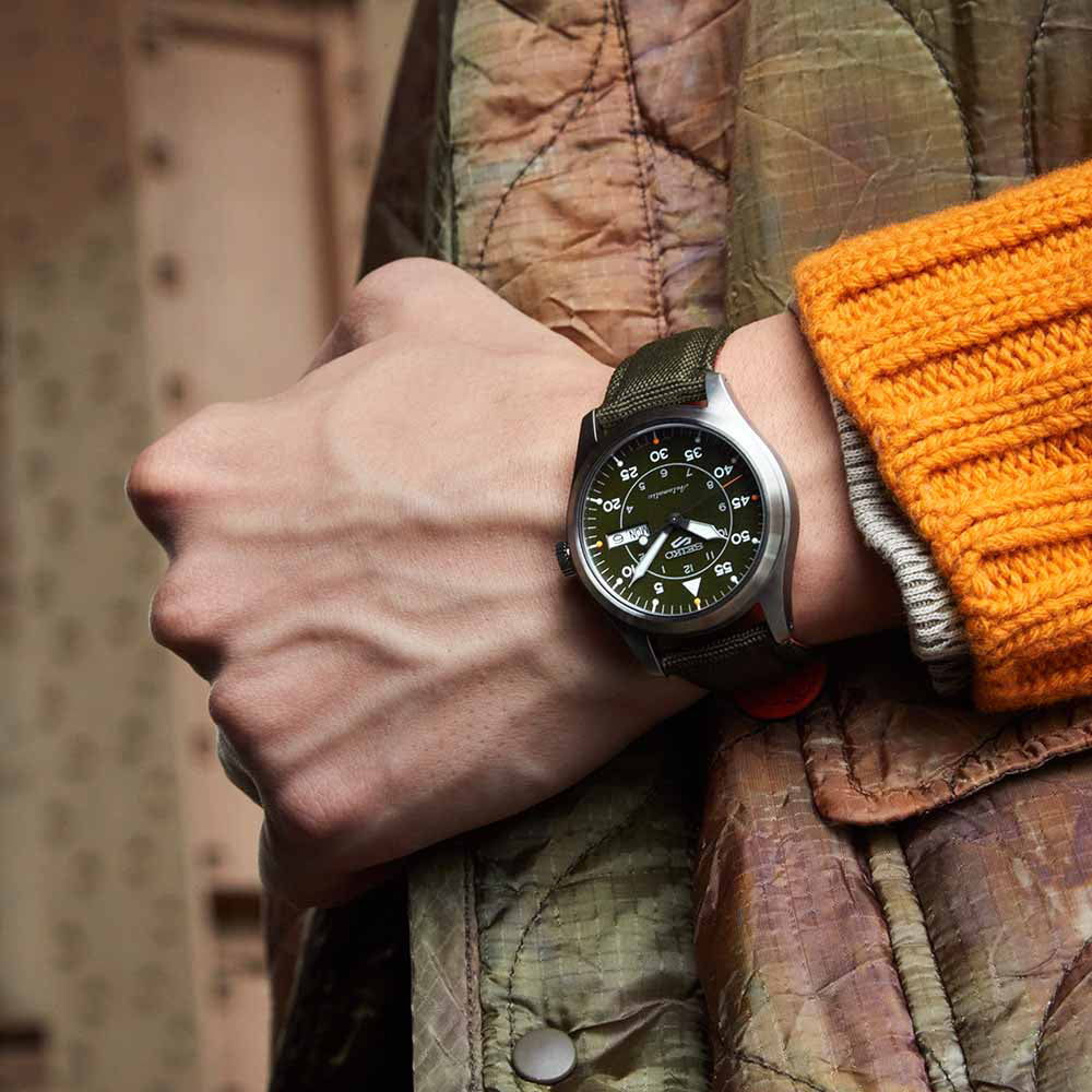 Een persoon die een Seiko 5 Sports herenhorloge SRPH29K1 met een zwarte wijzerplaat en witte cijfers draagt, draagt een beige gewatteerd jasje en oranje gebreide mouwen, wat zijn verfijnde look compleet maakt.