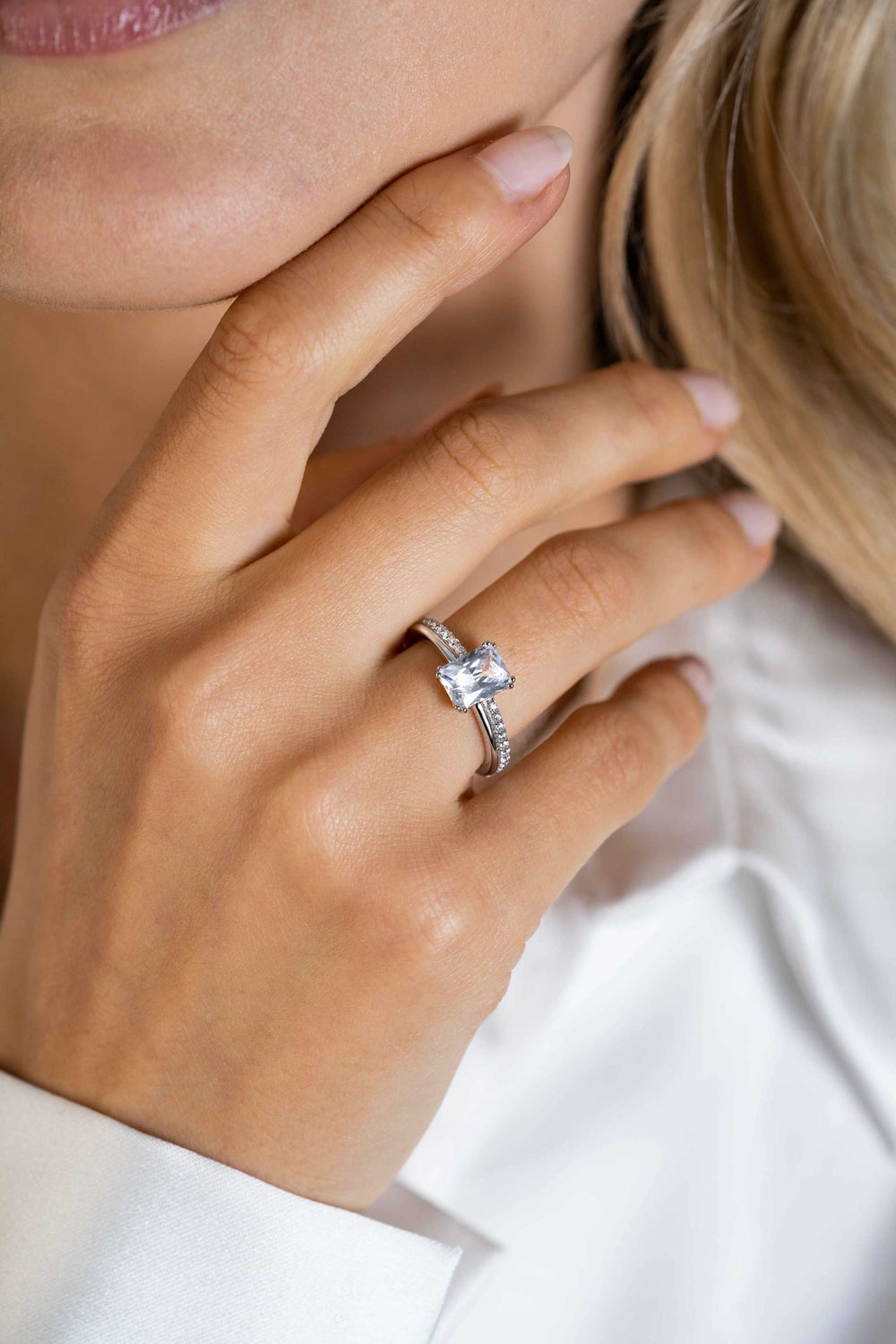 Een close-up van een vrouwenhand rustend op haar gezicht, met de Zinzi Ring Zilver Zir2392 met een witte zirkonia band en opvallende rechthoekige steen van Zinzi. Ze draagt een witte blouse en heeft netjes gemanicuurde nagels.