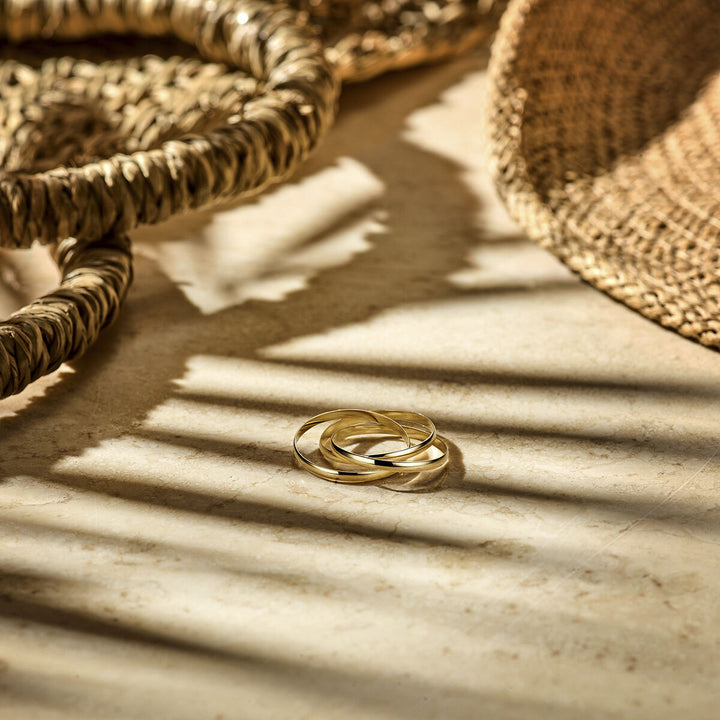 Drie Trinity ringen in 14 karaat geelgoud uit de Van der Kooij Collectie rusten op een licht stenen oppervlak, met gevlochten manden ernaast. De schaduwen van de manden werpen strepenpatronen op het warme, natuurlijke licht.