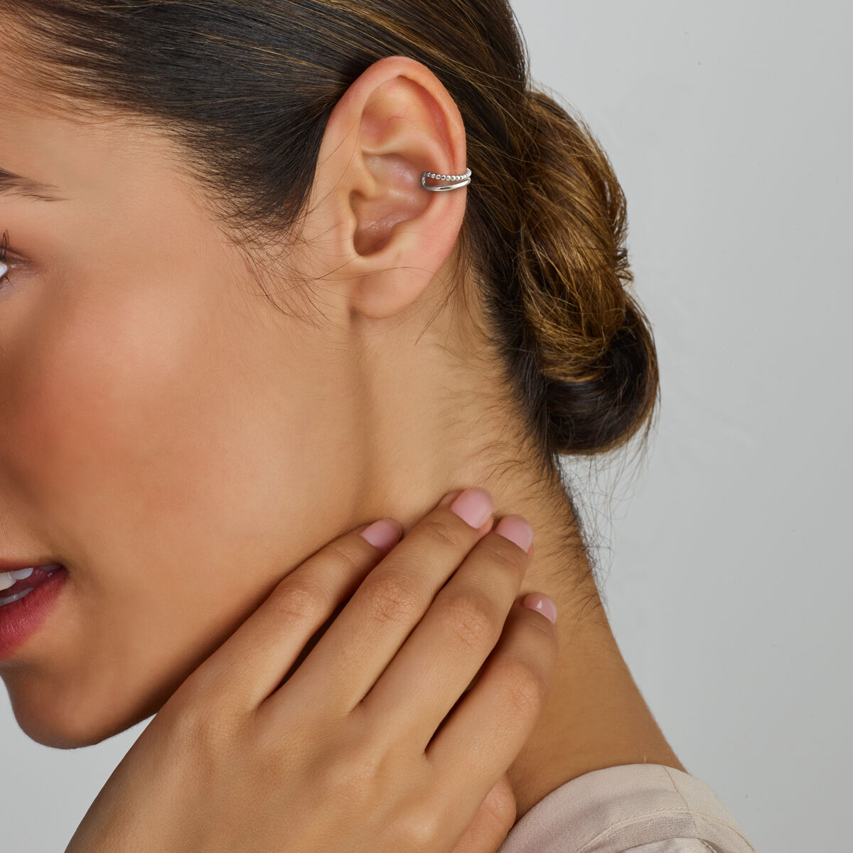 Een vrouw met donker haar in een lage knot draagt een Van der Kooij Collectie oorsteker zilver gerhodineerd aan haar linkeroor, haar gemanicuurde hand raakt haar nek. De effen lichtgrijze achtergrond accentueert de stijlvolle damesjuwelen.