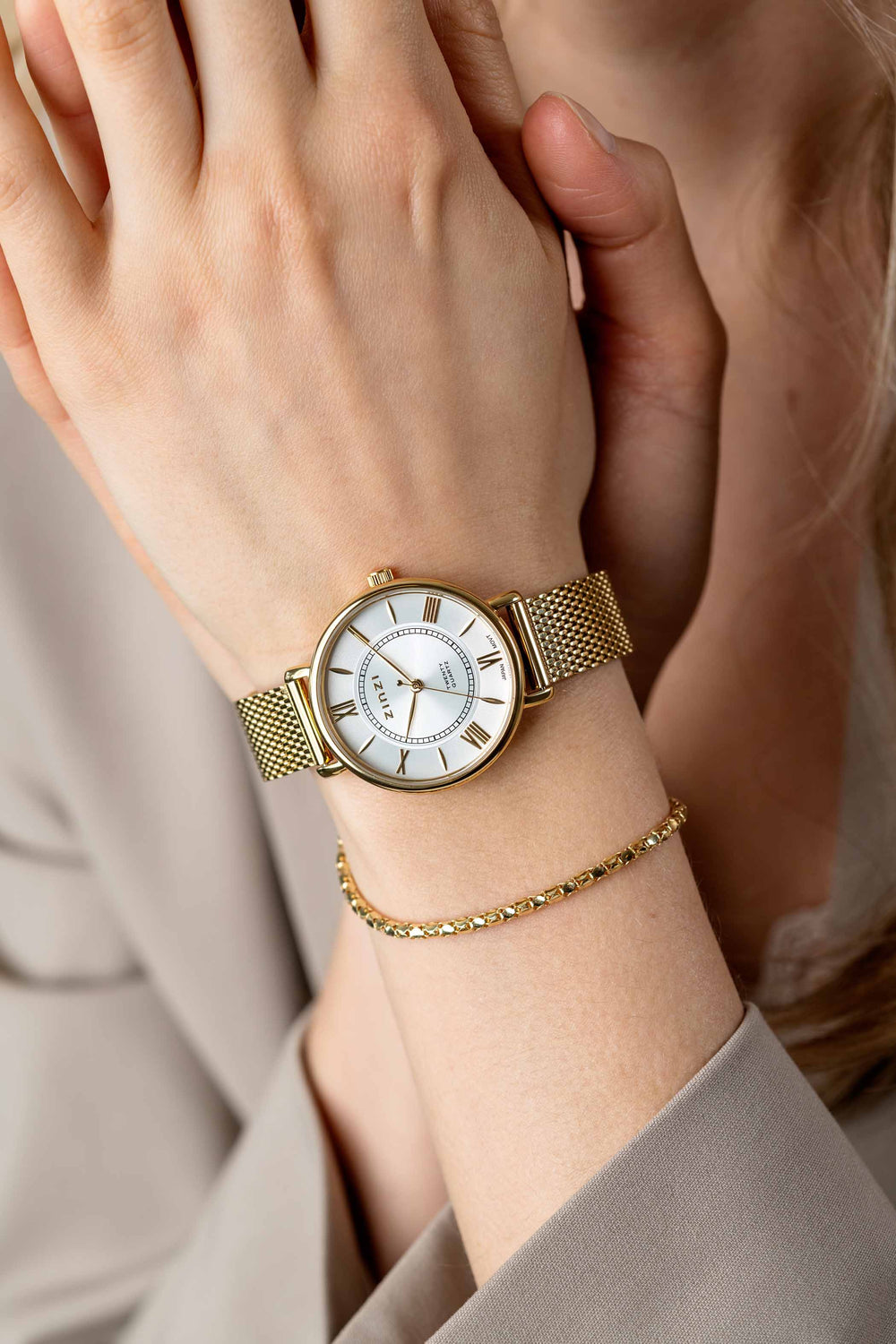 Een close-up toont een pols die de Zinzi Geelgoudkleurig Horloge Gold Plated Ziw2033 draagt met een band van goudgaas en een wijzerplaat met witte Romeinse cijfers, gestileerd met een fijne gouden armband terwijl de persoon zijn hand tegen zijn borst houdt.