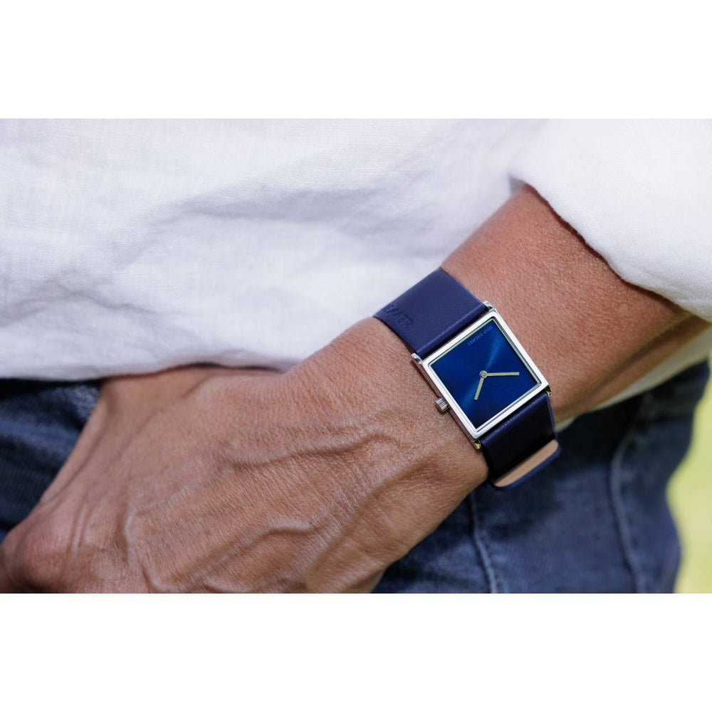 Close-up van iemand die een Rolf Cremer Horloge 508202 Groovy draagt, met een vierkant blauw quartzhorloge met bijpassende band, hand in zak van blauwe jeans. Ze zijn gekleed in een wit shirt, en tonen dit stuk uit de tijdloze collectie van het merk.
