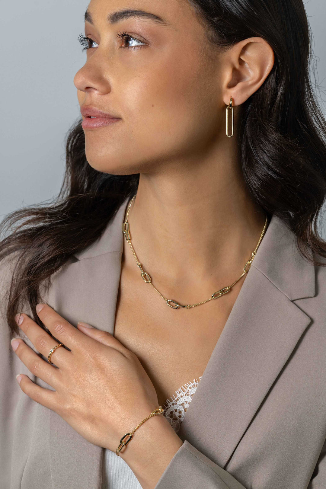Een vrouw in een lichtbeige blazer en wit kanten topje showt Zinzi Oorbedels Gold Plated Zich2416 G sieraden van Zinzi, waaronder een kettingketting, hoepeloorbellen, een ovaalvormige armband en een ring terwijl ze naar links kijkt.