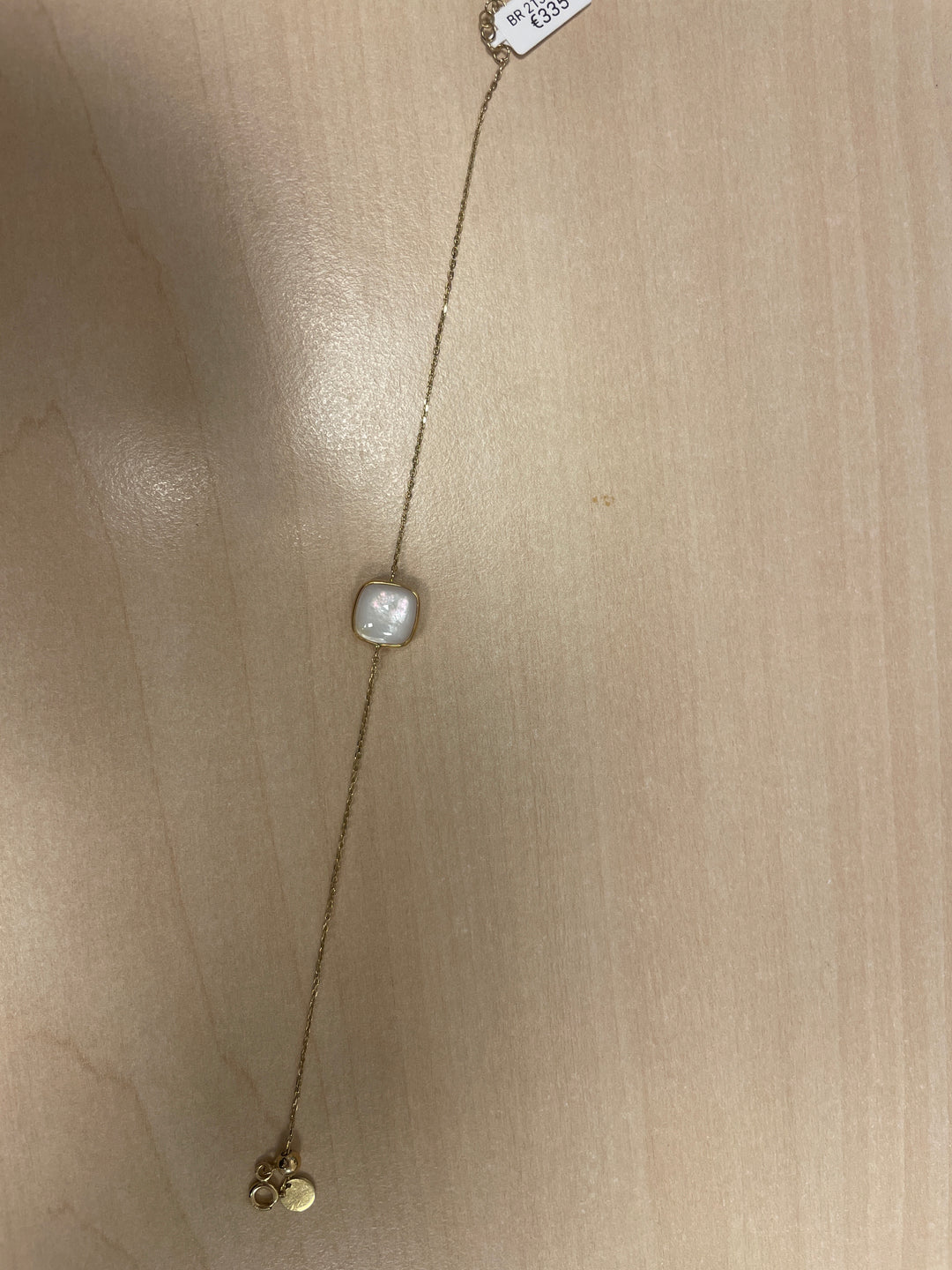 Op een licht houten oppervlak ligt een delicate Italo Bottene BR 2132-MOP gouden schakelarmband met een centraal vierkant parelachtig element en kleine gouden bedels aan één uiteinde. De prijs bedraagt £ 30,- bij de sluiting.