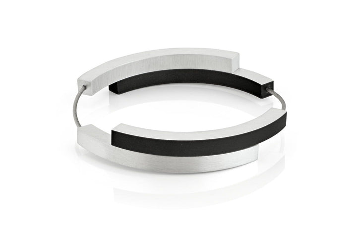 De Armband Vier bogen Zwart A32Z van CLIC Creations is een moderne, minimalistische bangle met afwisselende segmenten van zilver en zwart, verbonden door een dunne draad. Het vormt een halfopen cirkel voor een strakke, stijlvolle look. Handgemaakt in Nederland, het wordt weergegeven op een wit reflecterend oppervlak.