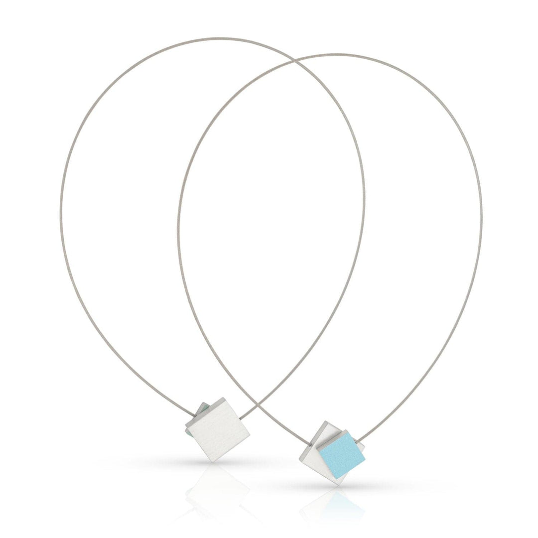 De Collier Vierkant met gekleurd vierkant C263B van CLIC Creations heeft minimalistische zilveren lussen met geometrische hangers: één lichtblauw vierkant en één effen zilver. Ze vormen een overlappende V en zijn vastgezet met een magnetische sluiting voor eenvoudig dragen.