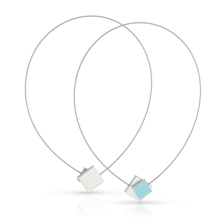 De Collier Vierkant met gekleurd vierkant C263B van CLIC Creations heeft minimalistische zilveren lussen met geometrische hangers: één lichtblauw vierkant en één effen zilver. Ze vormen een overlappende V en zijn vastgezet met een magnetische sluiting voor eenvoudig dragen.