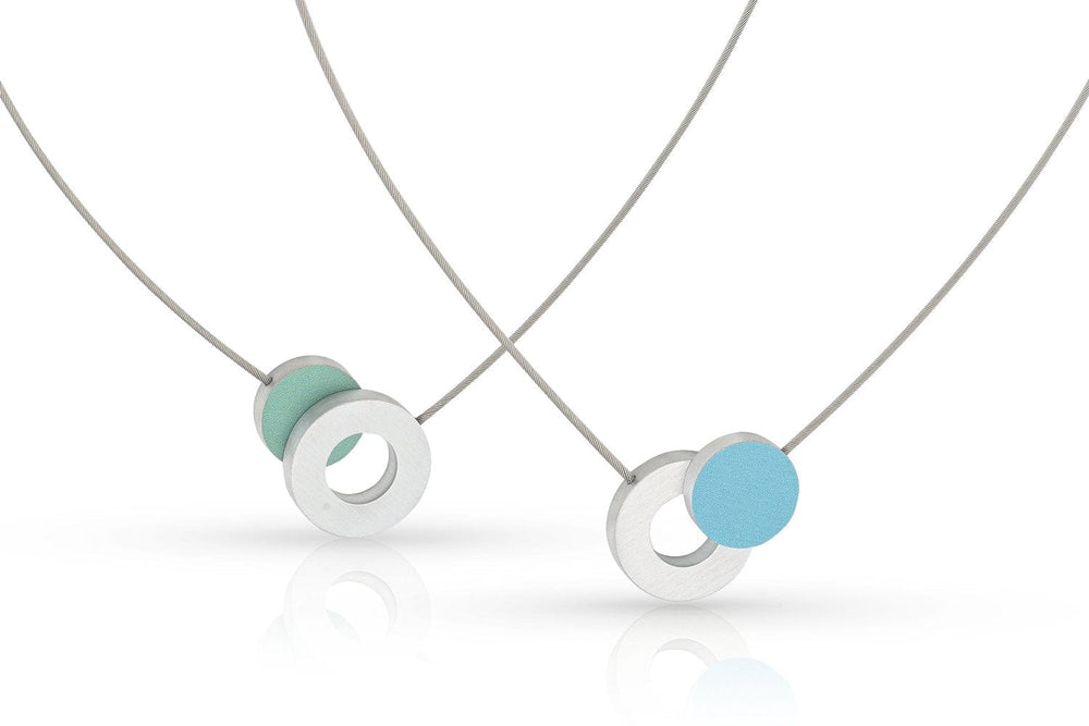 Twee minimalistische Collier Gekleurd rondje en een ring C265B van CLIC Creations hebben dunne kettingen met zilveren ringen en gekleurde cirkelvormige bedels, één blauw en één groen. Gekruist gerangschikt op een witte achtergrond, tonen deze kettingen handgemaakt in Nederland vakmanschap.
