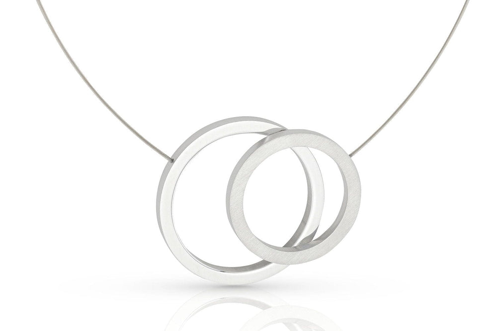 De CLIC Creations Collier grote ringen C267 heeft een modern design met twee in elkaar grijpende zilveren cirkels op een dunne ketting en een magnetische sluiting. De gladde, gepolijste ringen bieden elegantie tegen een effen witte achtergrond.