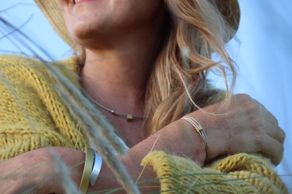Een vrouw met lang blond haar lacht warm, draagt een gele trui, beige hoed en sieraden. Haar hand op haar schouder benadrukt armbanden met magnetische sluiting. De scène is zacht wazig op de achtergrond. Aanbevolen product: Collier Klein Kubusje Rood C230R van CLIC Creations.
