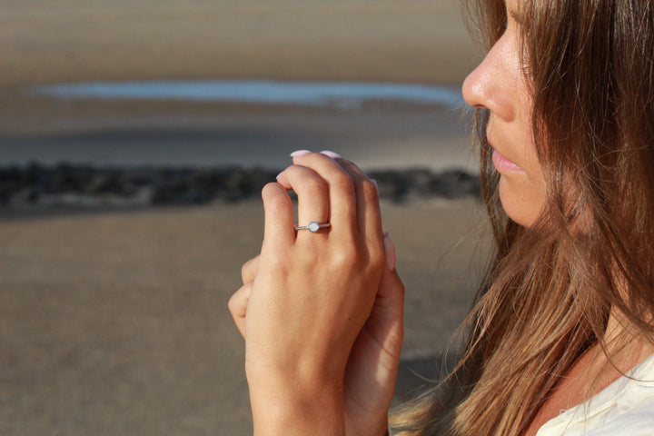 Een persoon met lang bruin haar houdt zijn handen bij zijn gezicht en toont de Isabelle | AsRing Zilver van See You aan zijn vinger. Hij staat op het strand, met op de achtergrond het glinsterende zand en water.
