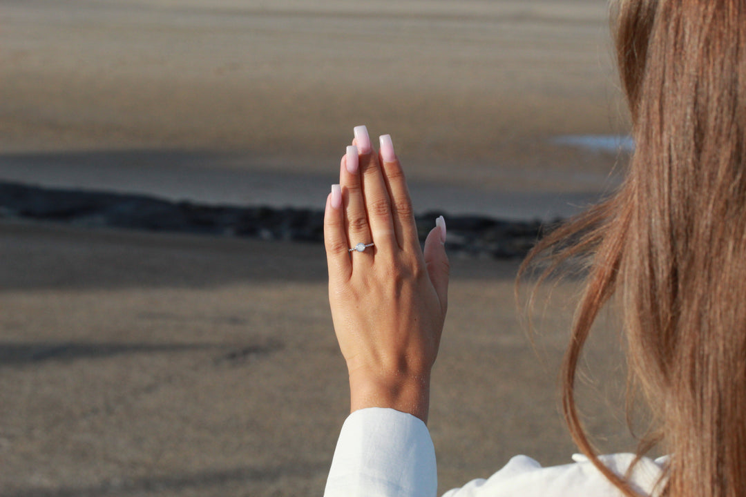 Een hand met lange, lichtroze gemanicuurde nagels en een Isabelle | AsRing Zilver van See You is opgeheven tegen een wazig zandstrand. Het individu draagt een wit shirt met lange mouwen.