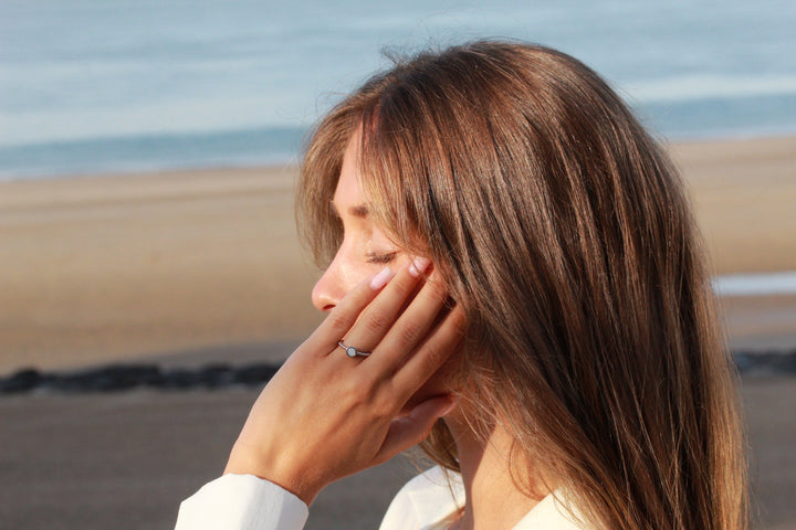 Een vrouw met lang bruin haar raakt haar gezicht aan en toont een See You Isabelle | AsRing Zilver aan haar vinger. Staand op een strand met de oceaan op de achtergrond, glinstert haar persoonlijke assieraad in de zon.