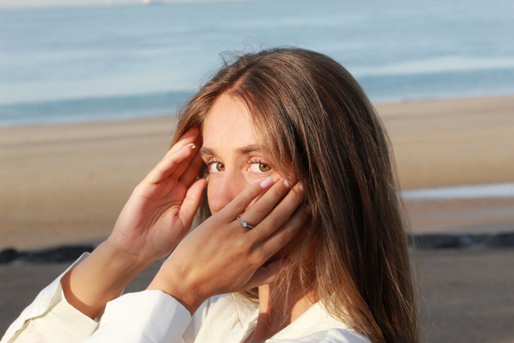 Een vrouw met lang bruin haar staat op een strand, met de oceaan achter haar. Ze kijkt in de camera, omlijst haar gezicht met haar handen terwijl ze een wit shirt draagt. Aan de ene kant toont ze de glinsterende Isabelle | AsRing Zilver van See You.