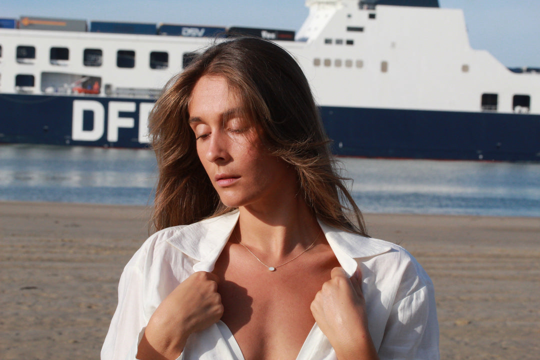 Op een zandstrand houdt een vrouw in een licht open wit shirt de kraag zachtjes vast met gesloten ogen. Om haar nek hangt de Luna | AsKetting Zilver van See You, zachtjes glinsterend in het licht, terwijl een grote veerboot achter haar over kalm water drijft.