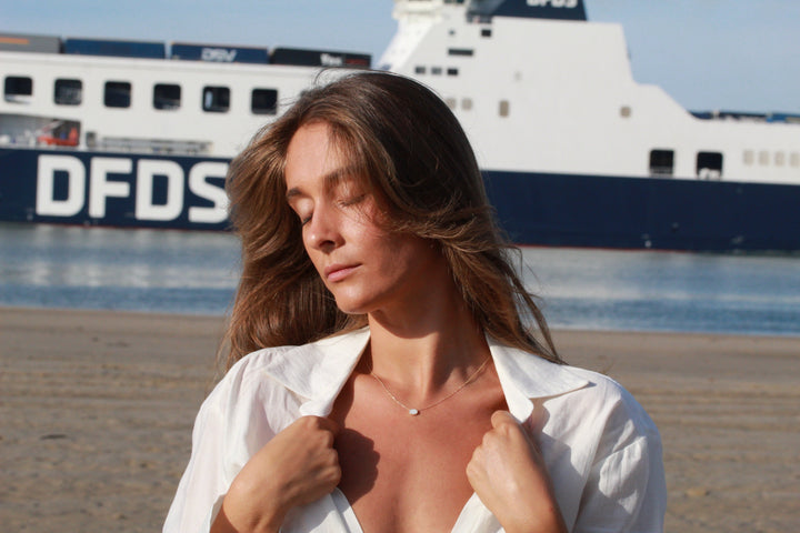 Een vrouw met lang haar staat op een strand, ogen dicht, draagt een wit shirt en een Luna | AsKetting Zilver van See You. Een grote veerboot met DFDS erop drijft vredig op de achtergrond tegen kalm water en een helderblauwe lucht.