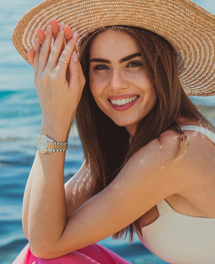 Een vrouw aan het water lacht naar de camera en draagt een breedgerande strooien hoed en een wit topje. Om haar pols draagt ze een stijlvol Jacques Du Manoir Inspiration 31 mm horloge.