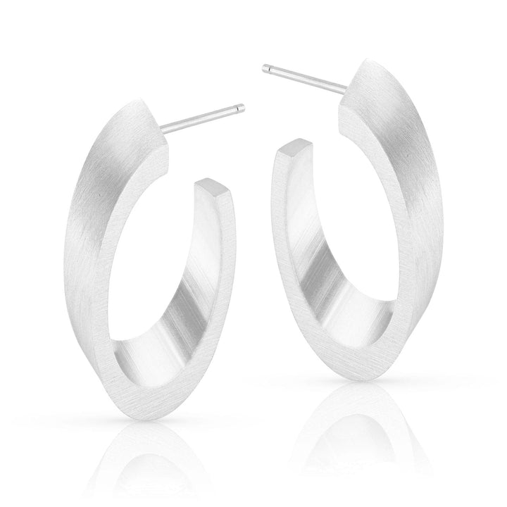 De Ovale Creolen O21 van CLIC Creations zijn zilveren creolen met een matte afwerking en open achterkant, met gladde, licht afgeplatte oppervlakken. Gemaakt van chirurgisch edelstaal, ze staan rechtop en worden weerspiegeld op een wit oppervlak.