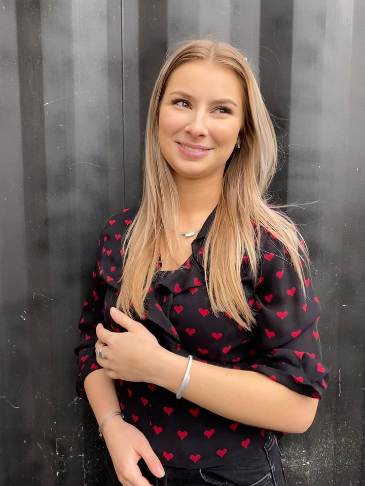 Een vrouw met lang blond haar lacht tegen een donkere, reflecterende achtergrond, gekleed in een zwart topje met rode hartjespatronen. Haar handen rusten lichtjes op haar middel. Ze showt het Oorsteker Schuin boogje O1 van CLIC Creations, dat doet denken aan handgemaakt in Nederland vakmanschap.