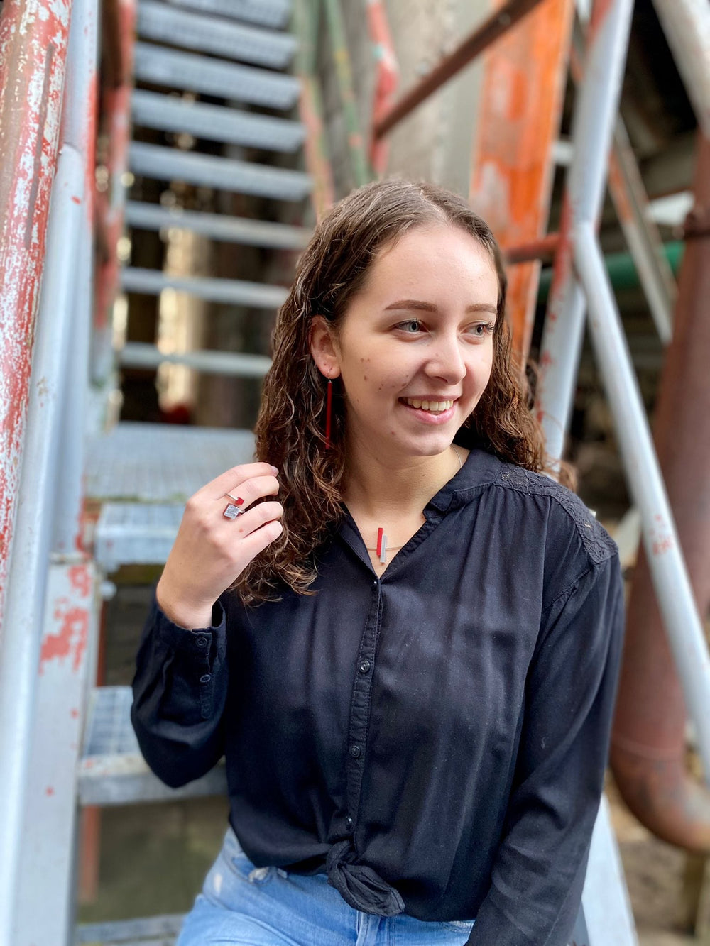 Een jonge vrouw met krullend haar, in een zwarte blouse en blauwe jeans, zit op een metalen trap. Ze glimlacht terwijl ze opzij kijkt en de Collier Eenvoudige staafjes Oranje C116O van CLIC Creations om haar nek aanraakt. De achtergrond toont versleten, veelkleurige leuningen.
