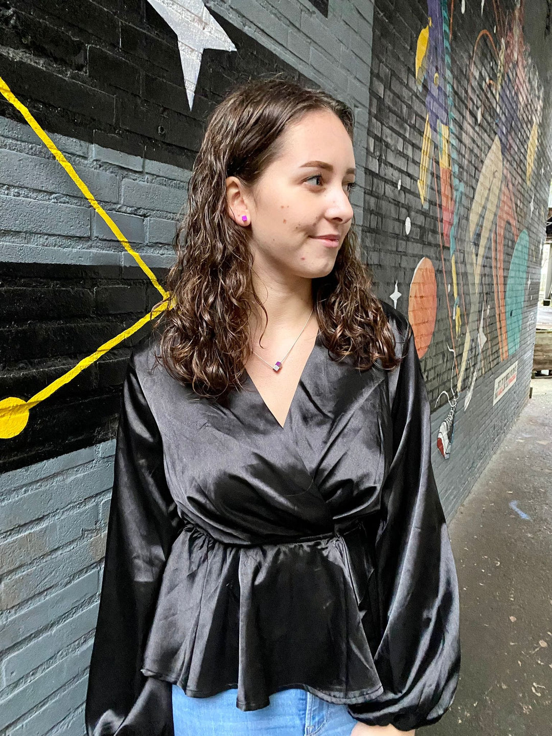 Een vrouw met krullend bruin haar, in een zwarte satijnen blouse en blauwe jeans, staat bij een met muurschildering bedekte bakstenen muur. De CLIC Creations Collier Twee kubusjes Oranje Paars met gouden draad CG205OP siert elegant haar nek, als aanvulling op de levendige geometrische vormen en sterren van de muurschildering.