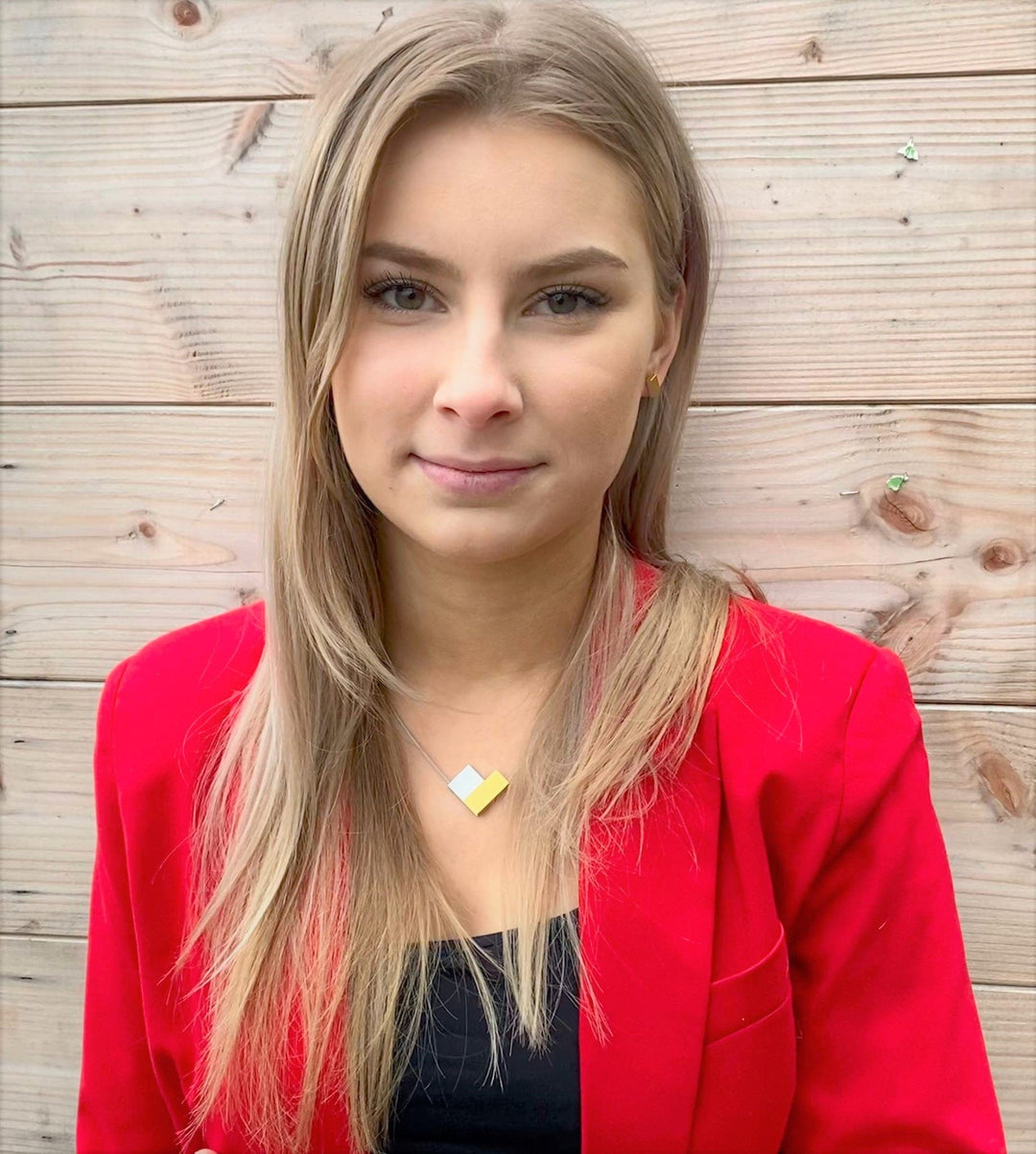 Een persoon met lang blond haar, gekleed in een rode blazer en een zwarte top, staat tegen een houten muur. Hun neutrale expressie wordt aangevuld door de CLIC Creations Collier V-vorm Blauw C150B, met een uniek ontwerp met ingewikkelde geometrische patronen en een handige magnetische sluiting.