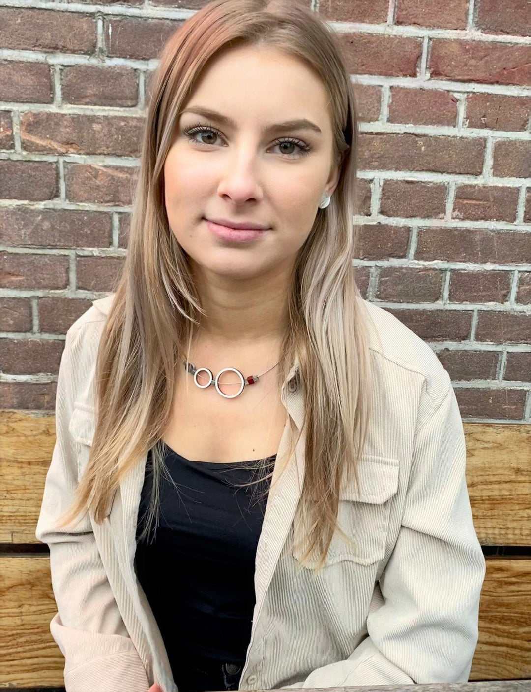 Een vrouw met lang blond haar zit tegen een bakstenen muur, met een beige shirt over een zwart topje. Haar CLIC Creations Collier Ringen Rood C186R ketting voegt elegantie toe aan haar look. Ze kijkt in de camera met een neutrale uitdrukking, wat subtiel het unieke ontwerp van haar accessoire benadrukt.