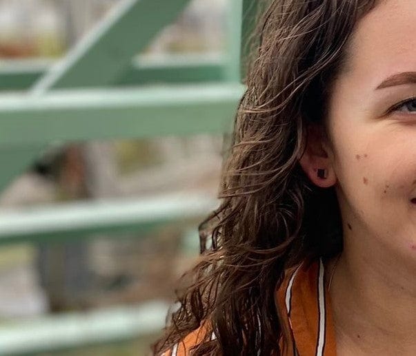 Close-up van een persoon met lang, golvend bruin haar, gekleed in een oranje en wit gestreepte top, met daarin Oorsieraad Kubusjes Paars O28P van CLIC Creations. De handgemaakte, hypoallergene oorstekers uit Nederland worden uitgelicht tegen een wazige achtergrond van een groene metalen structuur.