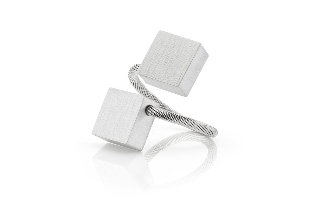 De Ring Vierkanten R6 van CLIC Creations is een minimalistisch metalen sculptuur, handgemaakt in Nederland. Het bestaat uit twee kubusvormige elementen die met elkaar verbonden zijn door gedraaide draad en heeft een zilveren afwerking, elegant gebalanceerd op een reflecterend oppervlak, wat lijkt op een perfect vervaardigde ring.