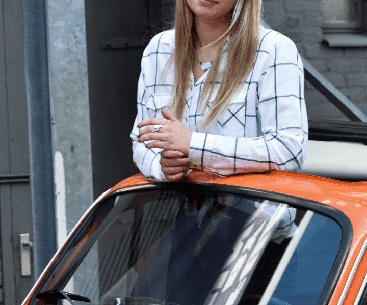 Een persoon met lang blond haar staat naast een oranje cabriolet, handgemaakt in Nederland, tegen een bakstenen muur en metalen trap. Ze dragen een wit shirt met een uniek design grid patroon van CLIC Creations.