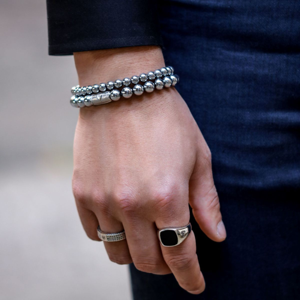 Een close-up van een hand met een zilveren kralenarmband en twee ringen van Rebel & Rose, één met een zwarte steen en één met een patroon, tegen een donkere jeans en een zwarte mouw.