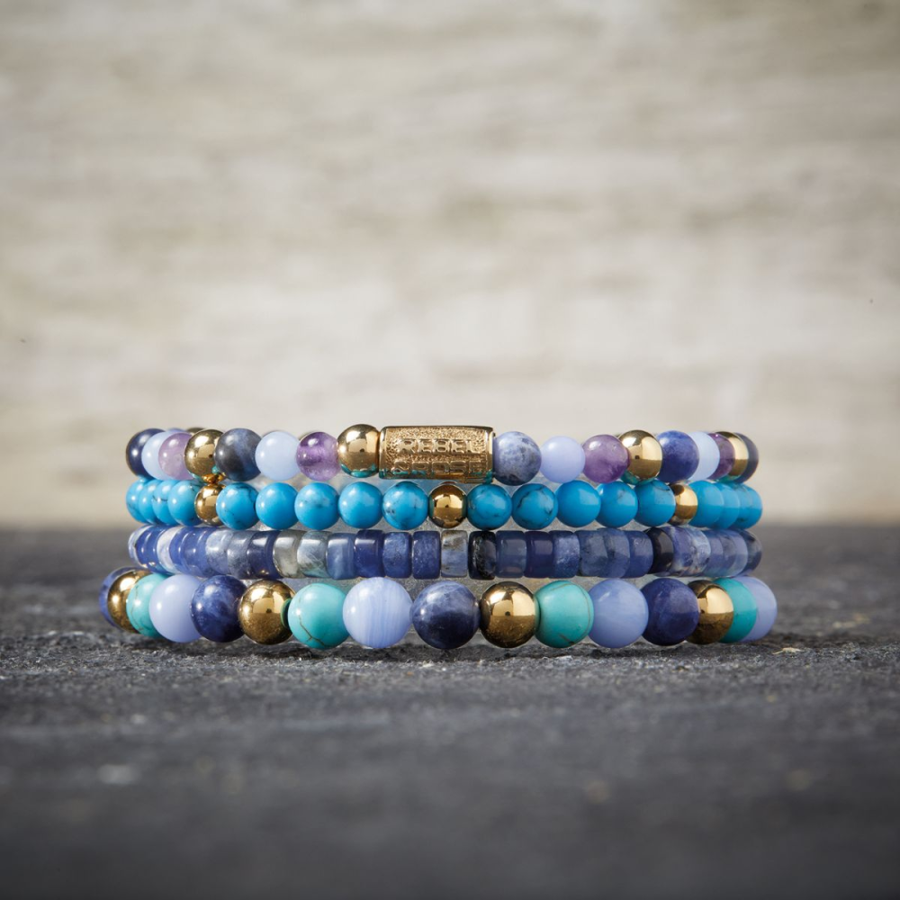 Een stapel kralenarmbanden met de Rebel & Rose Winter Blues - 18 kt geelgoud geïoniseerd 4mm Kralen Armband Dames wordt weergegeven op een grijs oppervlak met textuur en een vage lichte achtergrond.