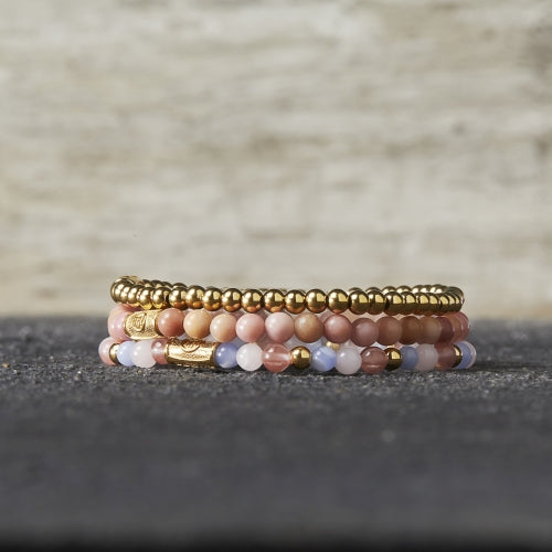 Een stapel kralenarmbanden van Rebel & Rose, waaronder de Rose Garden Gold - 4mm Kralen Armband Dames, heeft gouden, roze, lavendel en perzikkleurige kralen op een donker oppervlak - ideaal voor dames sieradenfans.