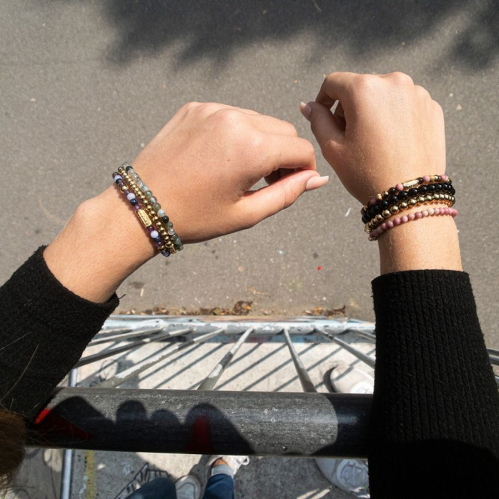 Twee handen met kralenarmbanden en een Rebel & Rose "Rose Garden Gold - 4mm - Kralen Armband Dames" zijn opgeheven over een metalen reling, hun schaduwen op het trottoir. De persoon in een zwarte top met lange mouwen benadrukt stijlvolle dames sieraden.