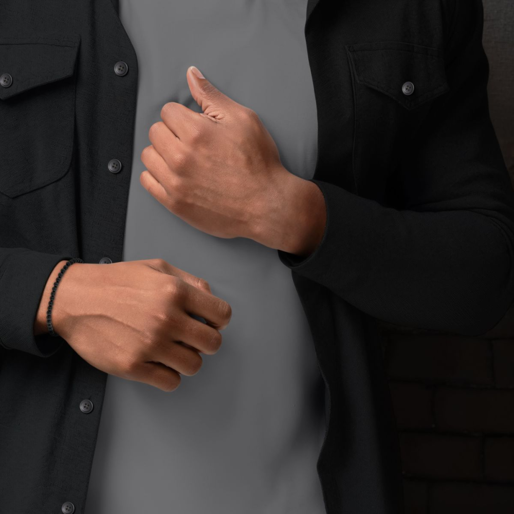 Een persoon in een zwart button-up shirt over een grijs t-shirt showt de Rebel & Rose Black Velvet - 4mm Kralen Armband Heren om één pols, met de handen en het bovenlichaam zichtbaar.