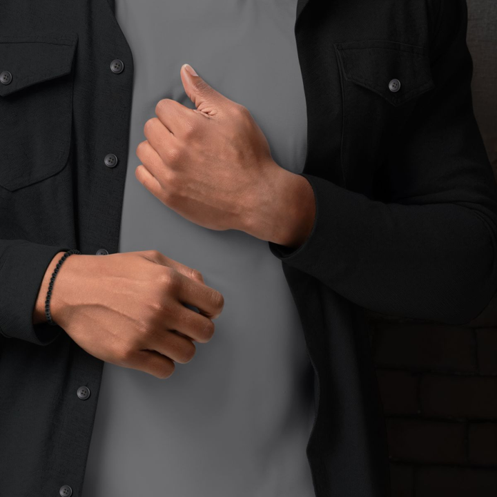 Een persoon in een zwart button-up shirt over een grijs t-shirt showt de Rebel & Rose Black Velvet - 4mm Kralen Armband Heren om één pols, met de handen en het bovenlichaam zichtbaar.