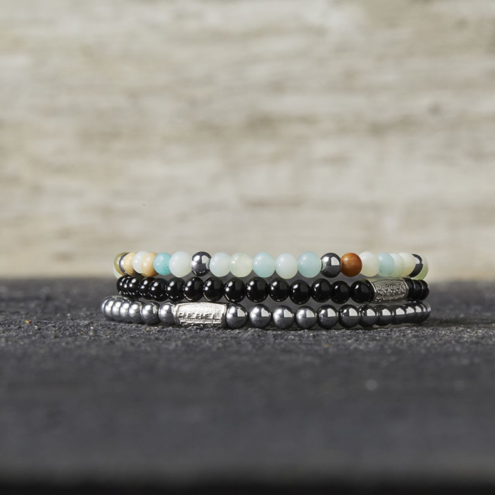 Drie Rebel & Rose Black Velvet 4mm kralen armband heren armbanden zijn gestapeld op een donker oppervlak, waarbij de bovenste veelkleurige kralen heeft en de anderen zwart en metallic grijs voor een elegante look.