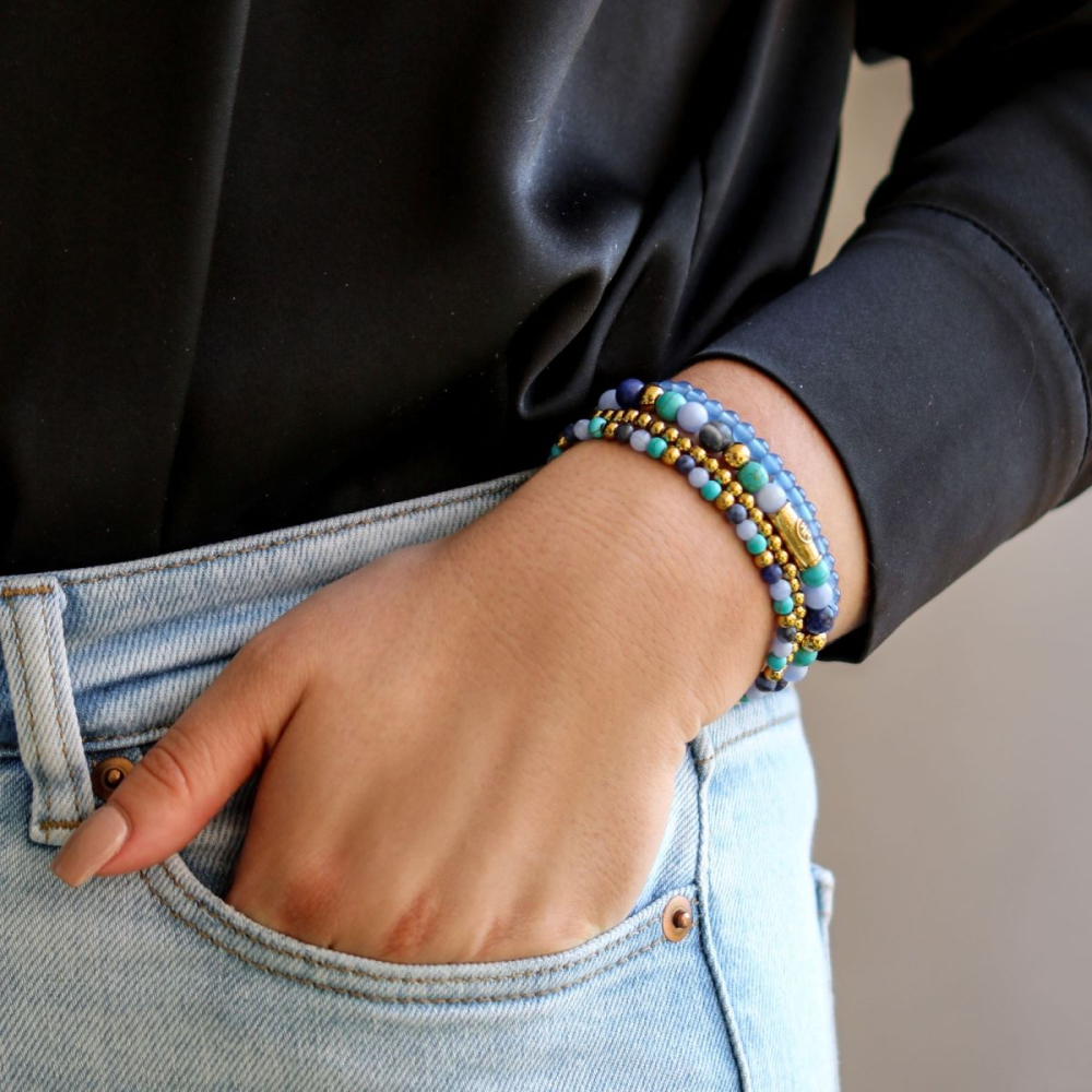 Iemand draagt een lichtblauwe jeans en een zwart overhemd en steekt zijn hand in zijn zak. De Rebel & Rose Winter Blues - 6mm Kralen Armband Dames zorgt voor een stijlvol sieraad en een vleugje winter blues elegantie.