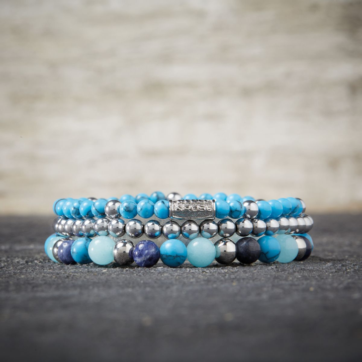 Een stapeltje van drie Rebel & Rose Blue Summer Vibes - 6 mm Kralen Armband Dames, met turquoise en sodalietkralen met een metallic label, weergegeven op een donker oppervlak - perfect voor een stijlvolle blauwe armbandlook.