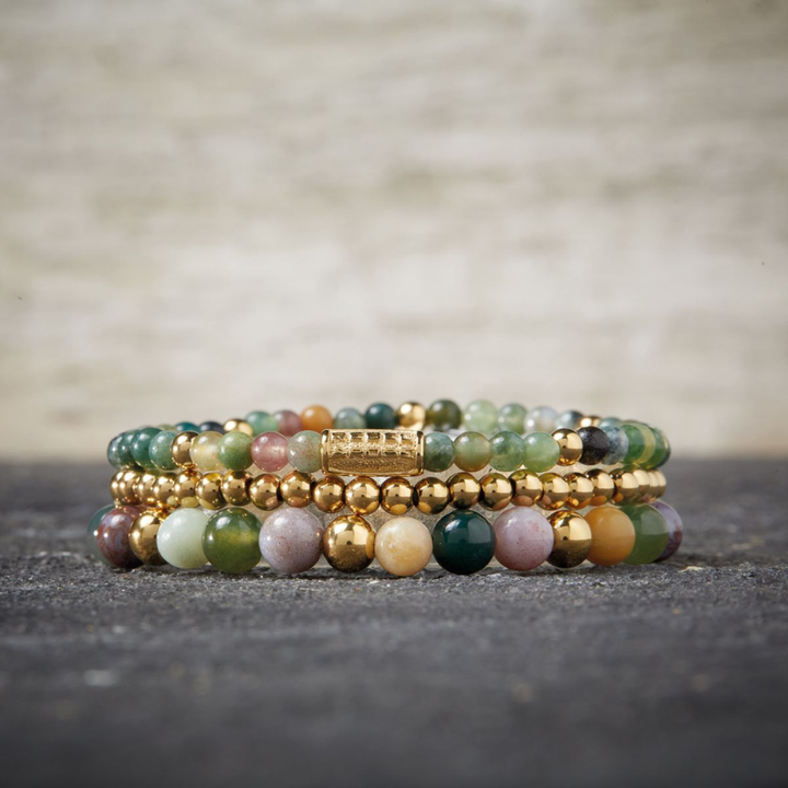 Een stapeltje van vier kralenarmbanden, met de Spring Shine armband van Rebel & Rose-18 kt geelgoud geïoniseerd, 6 mm kralen-displayed op een donker gestructureerd oppervlak met een neutrale achtergrond.