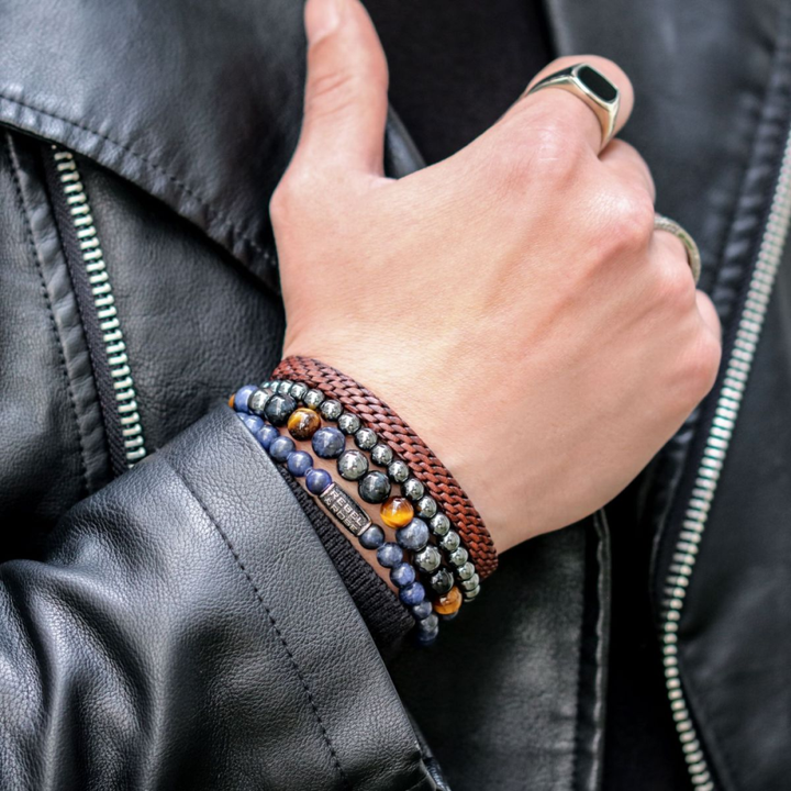 Een persoon in een zwart leren jack toont zijn hand met verschillende kralenarmbanden, waaronder de Rebel & Rose Grey Silver Shine DV - 6mm Kralen Armband Heren, plus een bruin gevlochten band en een dikke zilveren ring.