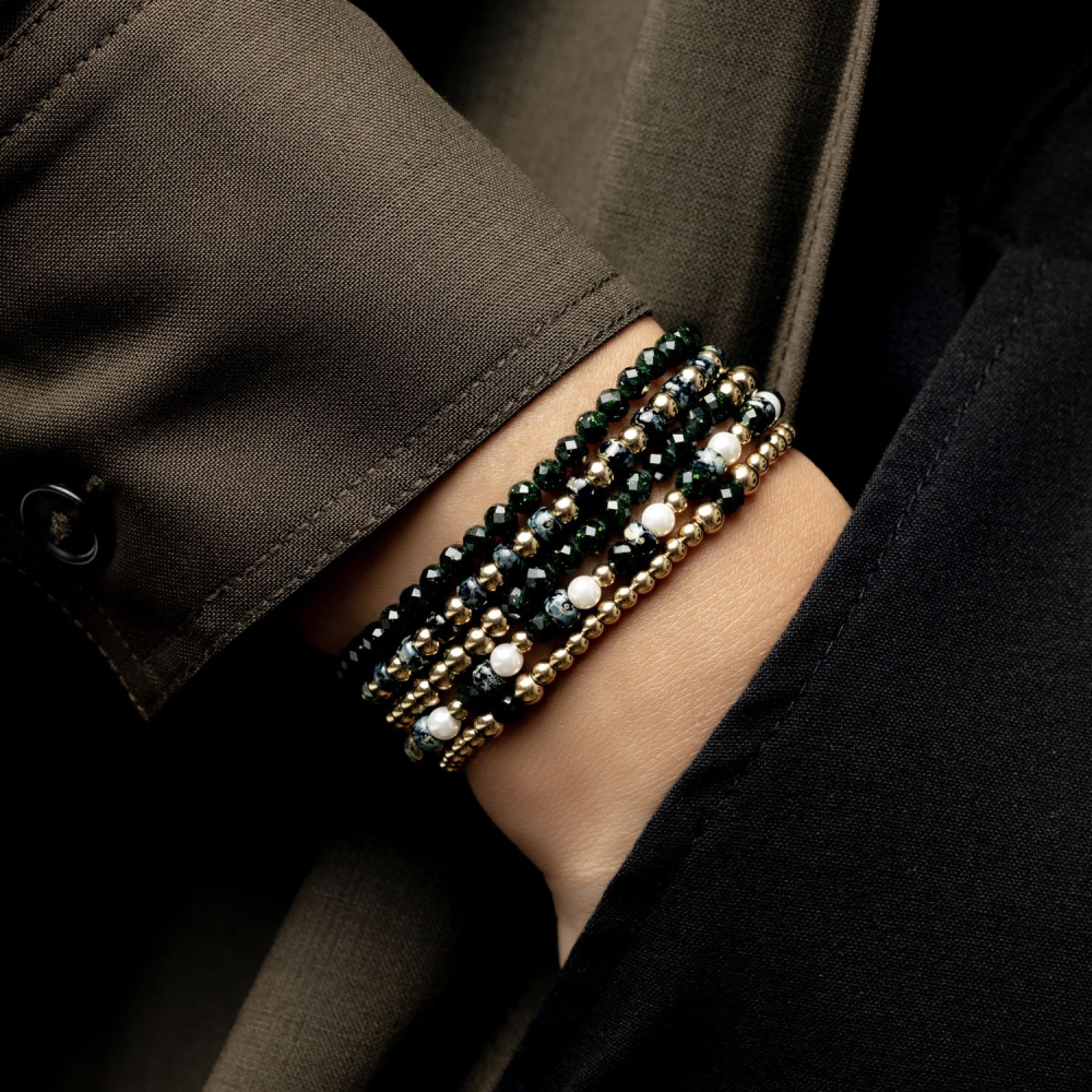 Een close-up van een pols met Rebel & Rose’s Green Glam Rocks Party - 4mm Kralen Armband Dames, met groene kralen en andere armbanden, gestyled over de mouw van een donker, formeel jasje.