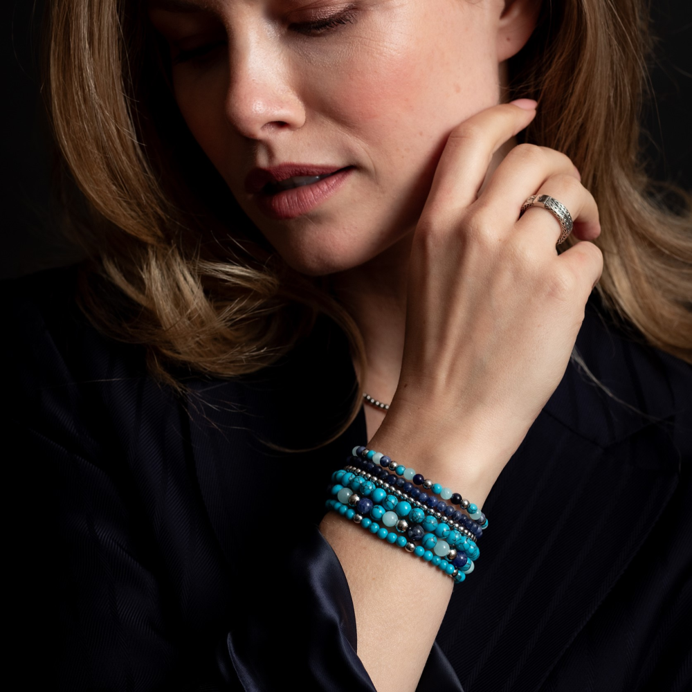 Een vrouw met golvend blond haar, gekleed in donkere kleding, staart naar beneden terwijl ze haar gezicht aanraakt, met de Blue Summer Vibes - 6mm Kralen Armband Dames van Rebel & Rose en een zilveren ring tegen een donkere achtergrond.