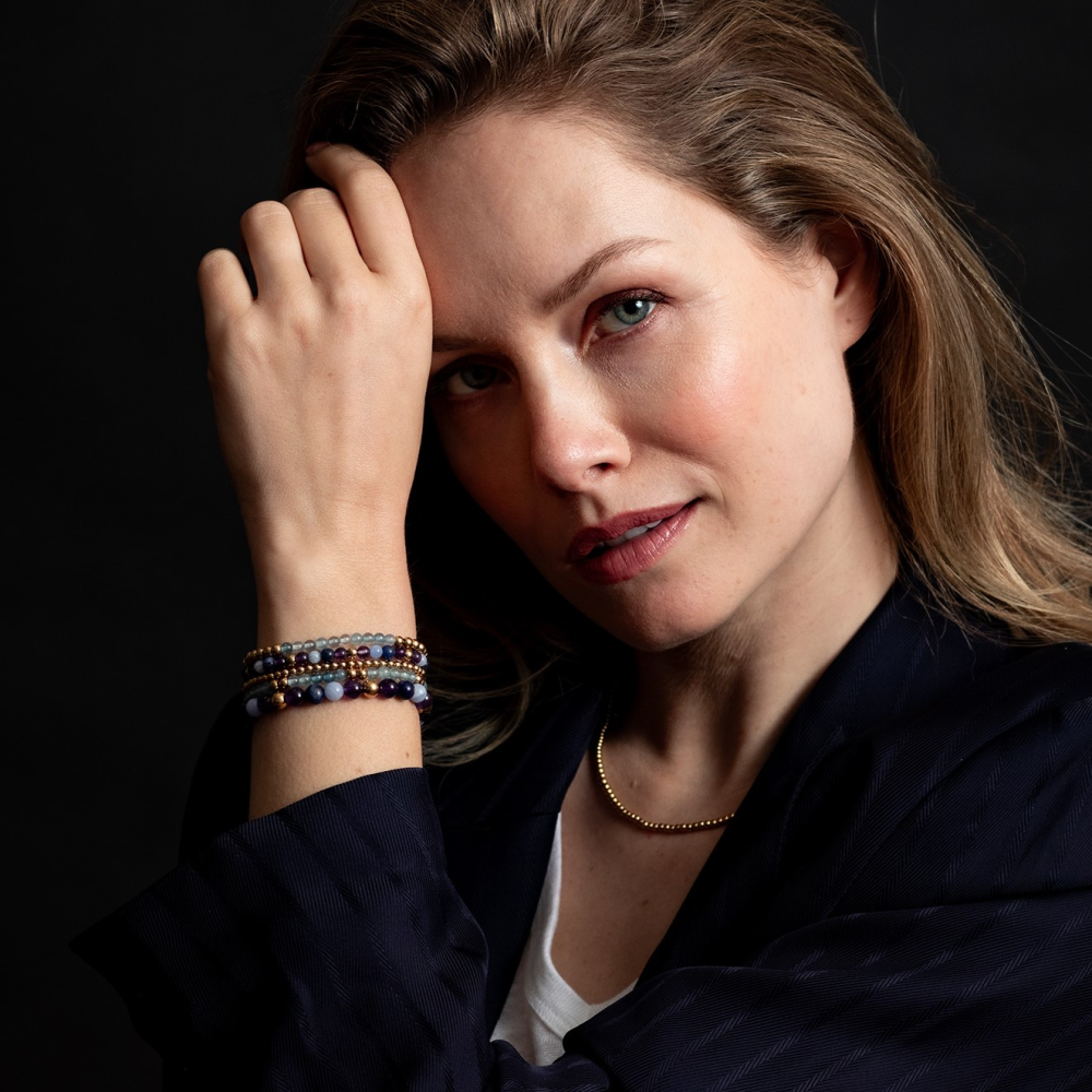 Een vrouw met lichtbruin haar draagt een donkere blazer, wit overhemd en de Rebel & Rose "Are Violets Blue?" armband. 18 kt geelgouden geïoniseerde 6mm kralenarmband. Ze kijkt naar de camera met haar hand op haar voorhoofd tegen een donkere achtergrond.