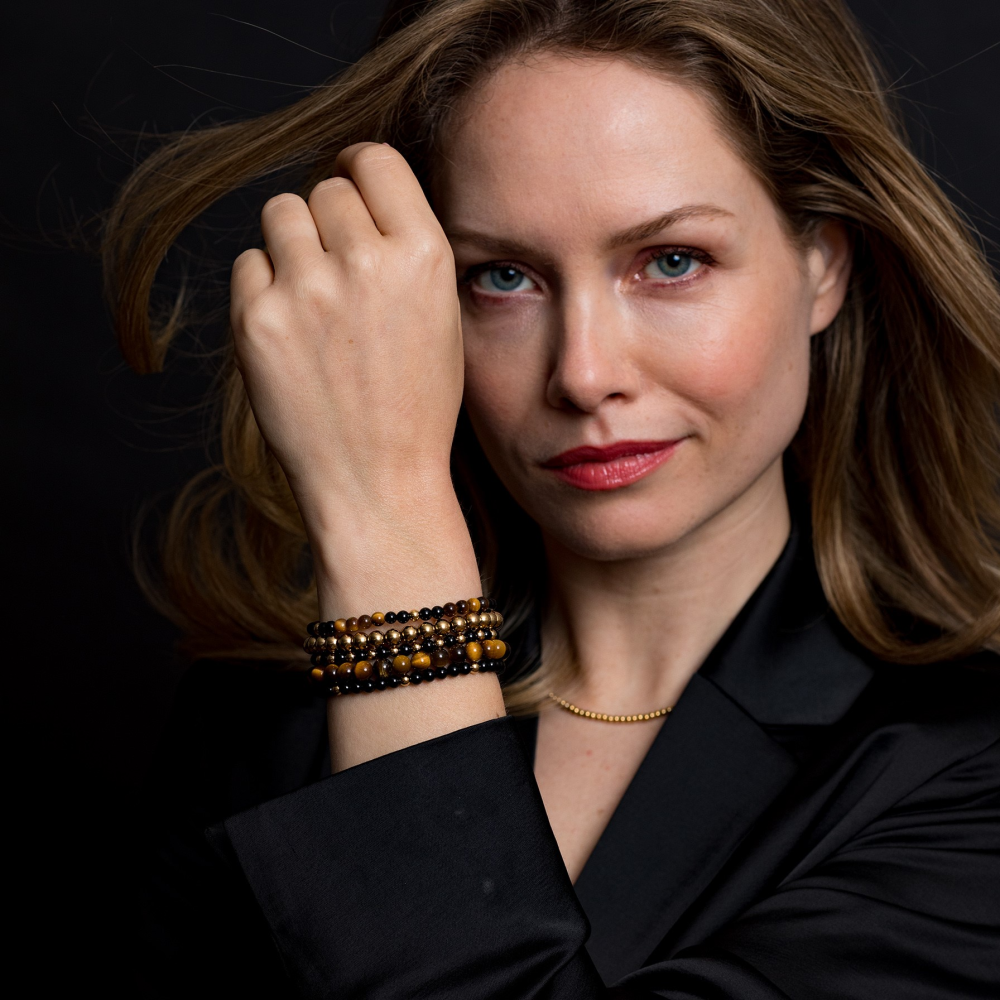 Een vrouw met lichtbruin haar en rode lippenstift poseert tegen een zwarte achtergrond. Ze draagt een zwart jasje, een gouden ketting en de Rebel & Rose Tiger Lily Gold - 6mm Kralen Armband Dames om haar pols, die ze bij haar gezicht houdt.