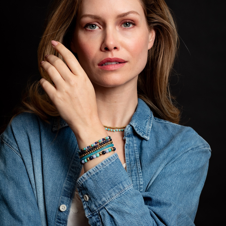 Een vrouw met lang lichtbruin haar in een spijkerhemd steekt haar linkerhand op en toont de Rebel & Rose Winter Blues - 6mm Kralen Armband Dames met turquoise agaat sodaliet kralen, plus kleurrijke armbanden en een gouden ketting tegen een donkere achtergrond.