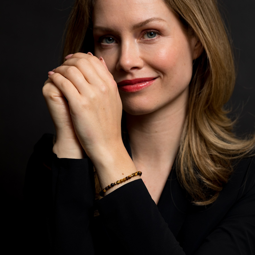 Een vrouw met lichtbruin haar, in een zwart topje en een Rebel & Rose Tiger Lily - 18 kt geel goud geïoniseerd 4mm Kralen Armband Dames, glimlacht zachtjes met haar handen tegen een donkere achtergrond.