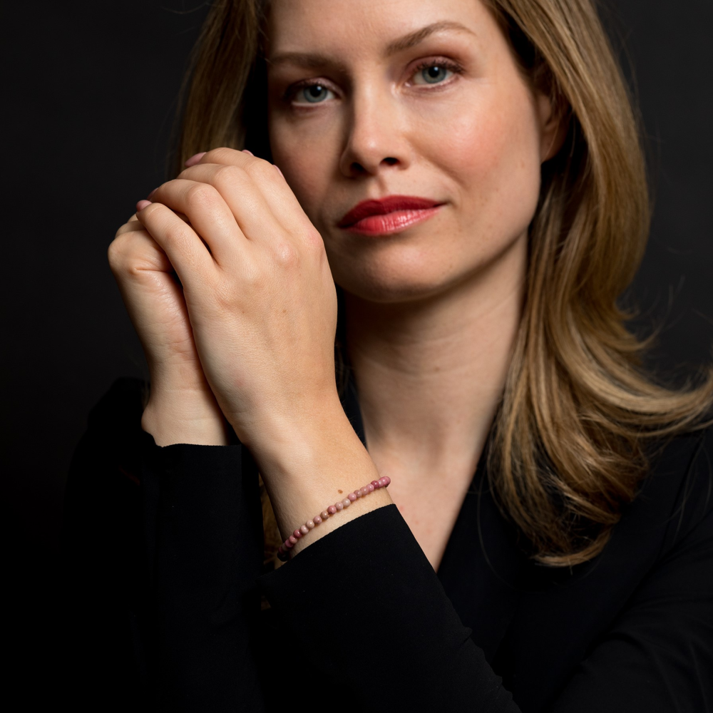 Een vrouw met lichtbruin haar en rode lippenstift poseert tegen een donkere achtergrond, draagt een zwart topje en de Rebel & Rose Rose Garden Gold 4mm kralen armband dames - haar handen zachtjes bij haar gezicht geklemd voor een verfijnde look.