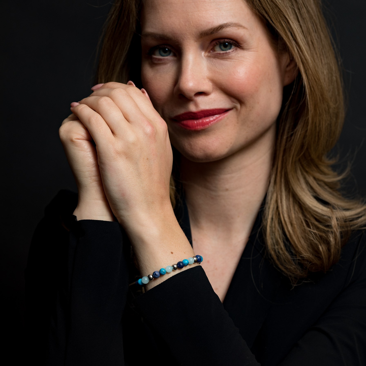 Een vrouw met lichtbruin haar glimlacht zachtjes, draagt een zwart topje en de Rebel & Rose Blue Summer Vibes - 6mm Kralen Armband Dames met sodalietkralen, haar handen zachtjes geklemd tegen een donkere achtergrond.